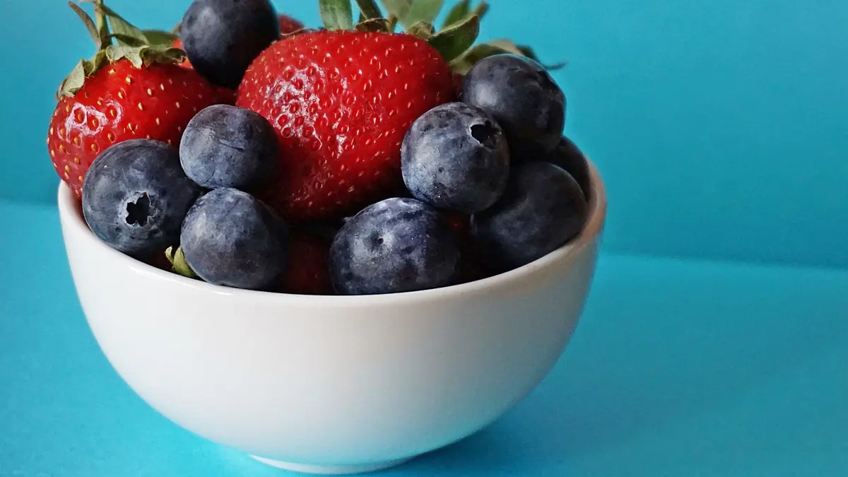Un bol blanco con fresas y arándanos sobre un fondo turquesa