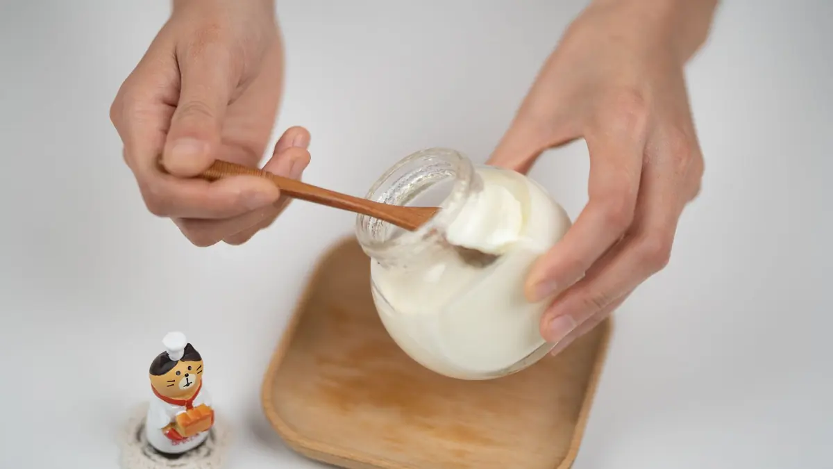Unas manos sacan con una cuchara de madera yogur natural de un frasco de vidrio, sobre una bandeja de madera