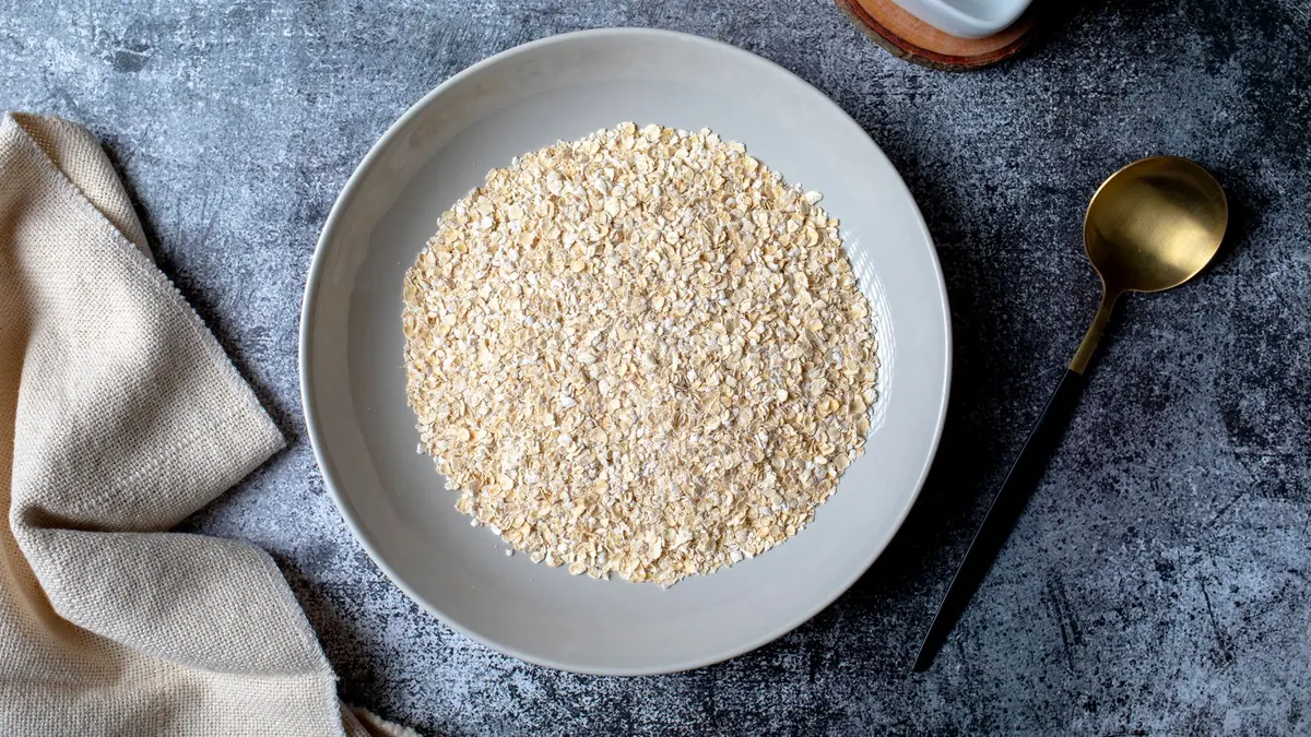 Avena en hojuelas en un plato hondo gris, con una cuchara dorada y un paño de lino al lado, sobre una mesa de piedra oscura