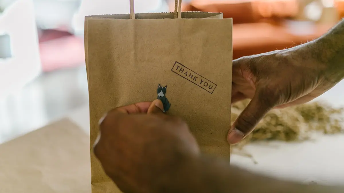 Una mano coloca una pequeña figura de un perro sobre una bolsa de papel kraft con un sello que dice Thank you