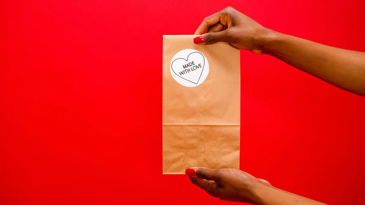 Dos manos sostienen una bolsa de papel kraft cerrada, con un sello redondo blanco con un corazón y las palabras MADE WITH LOVE, sobre un fondo rojo