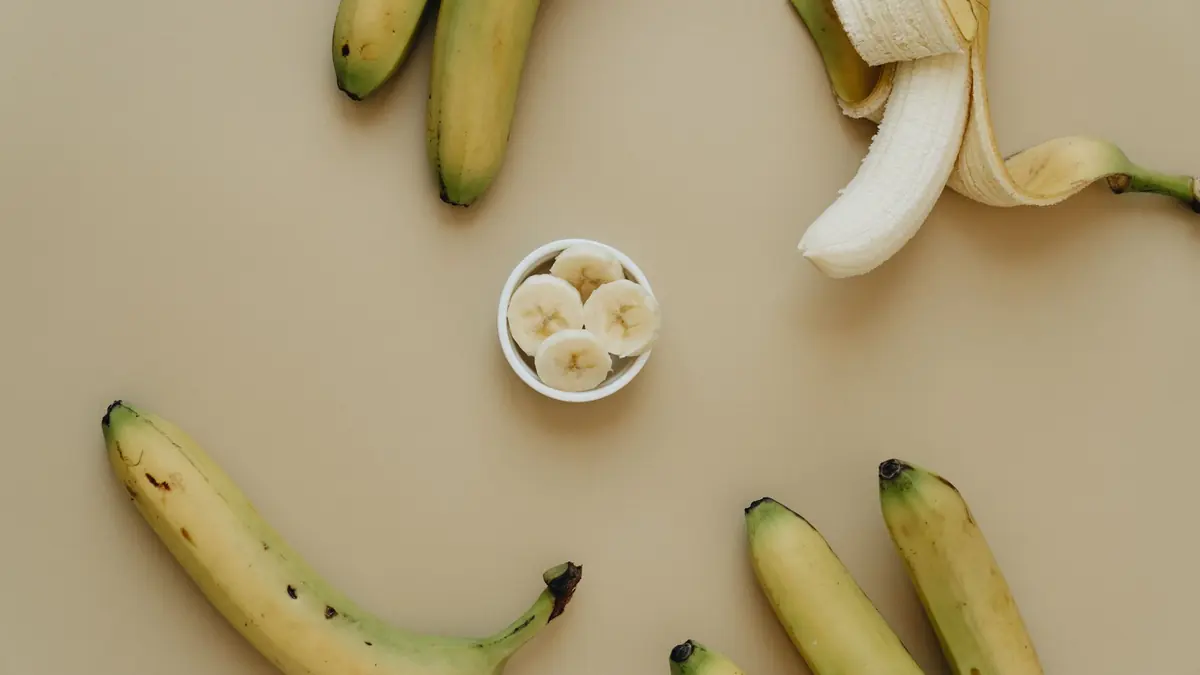 Plátanos sobre un fondo beige, uno con la cáscara abierta, y rodajas de plátano en un cuenco pequeño