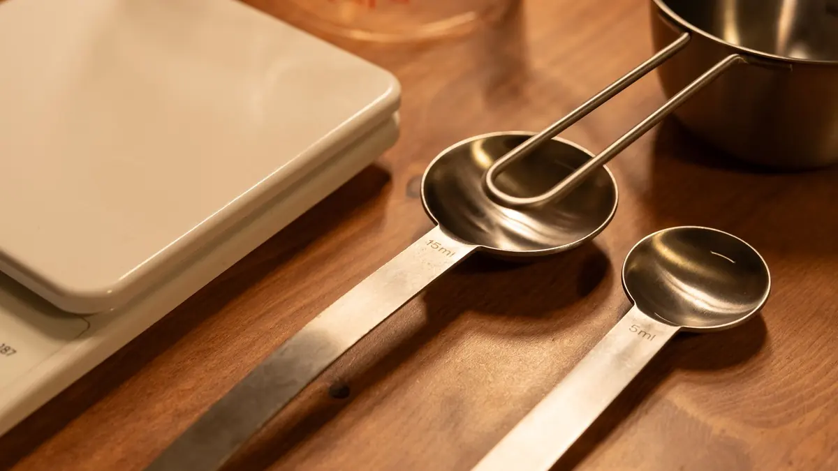 Una balanza de cocina digital, dos cucharas medidoras de acero de 15 y 5 ml y una taza medidora sobre una mesa de madera