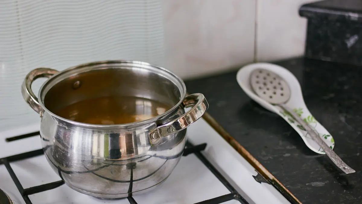 Una olla de acero inoxidable con un líquido dorado sobre una cocina a gas, con una espumadera apoyada al lado