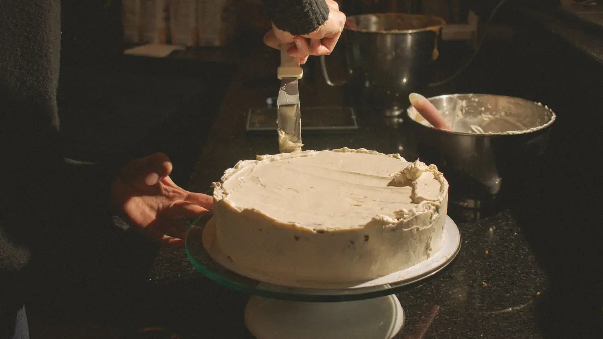 Una persona alisa con una espátula el betún blanco de un pastel para personas sobre un plato giratorio, en una cocina con poca luz