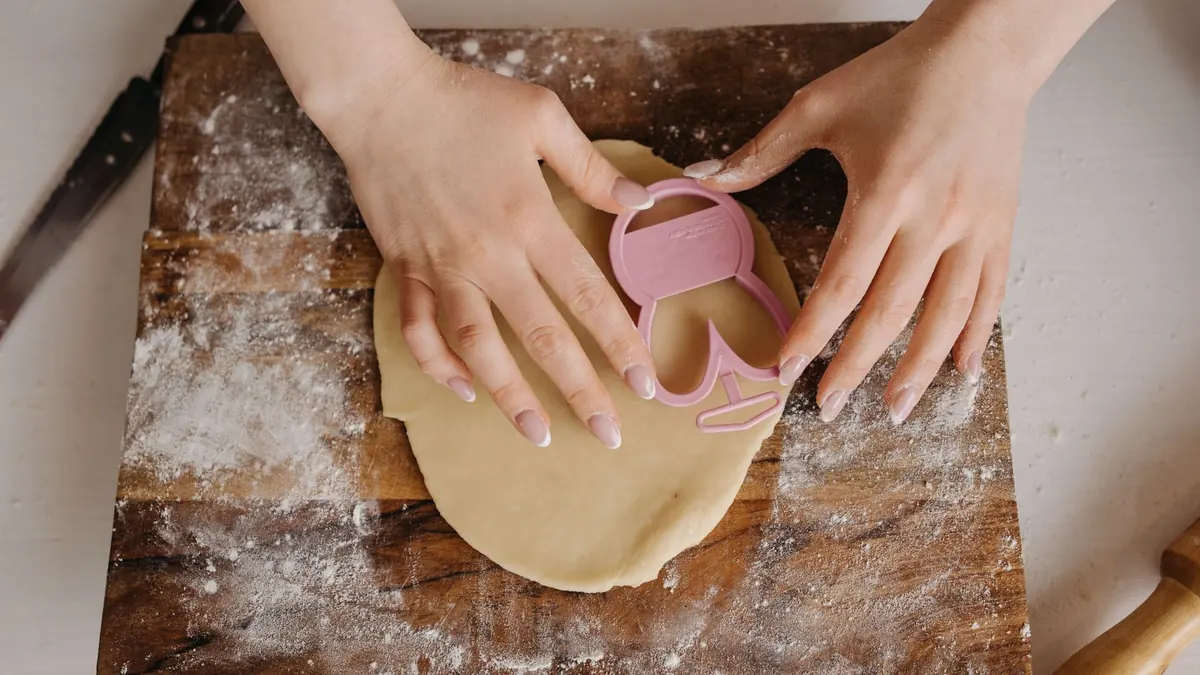 Manos que cortan una masa estirada con un cortador rosa sobre una tabla de madera con harina