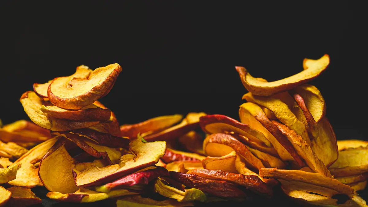 Un montón de rodajas de manzana deshidratada, con la cáscara roja y el centro dorado, sobre un fondo negro