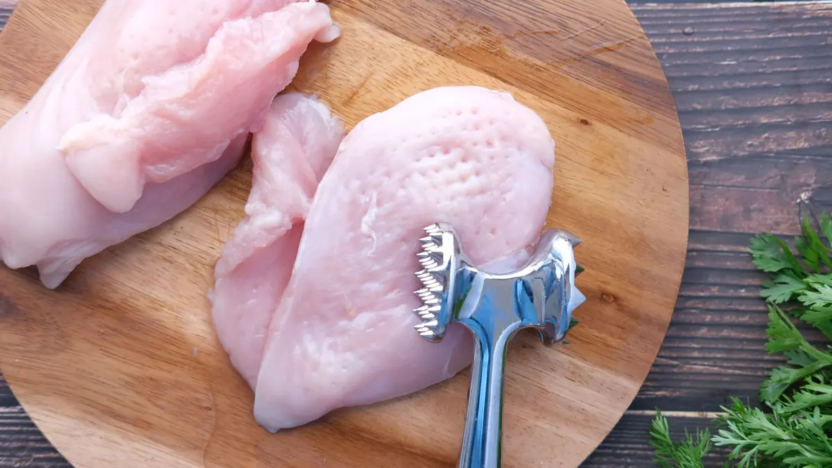 Dos pechugas de pollo crudas sobre una tabla de madera redonda, con un mazo para carne con dientes apoyado sobre una de ellas y unas hojas de perejil a la derecha